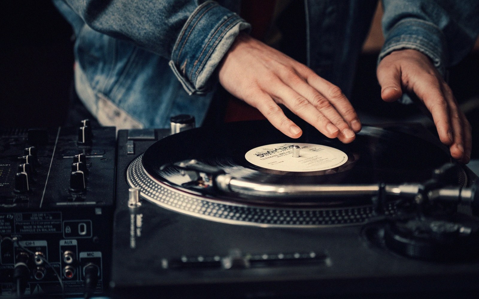 A DJ's hands rest on a spinning vinyl record beside a mixer